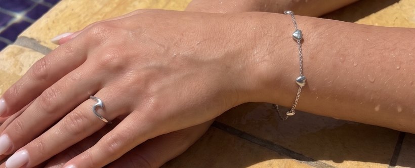 Mãos com anel de onda e pulseira de corações em inox à beira da piscina